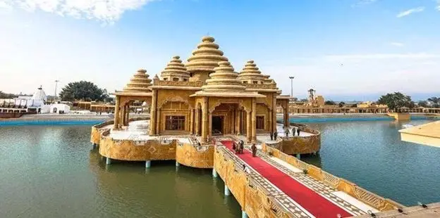 Hindu temple complex on a small island, connected by a red carpet bridge over calm water, with tan stone buildings and tiered domes.